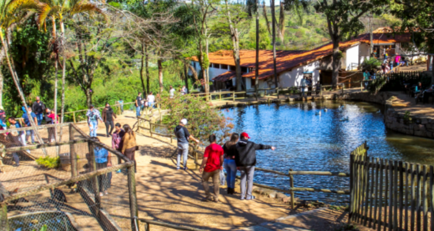 Vista da Fazenda do Chocolate em Itu, mostrando visitantes caminhando ao redor do lago, cercados por vegetação, animais e estruturas rústicas típicas do local. Collorau